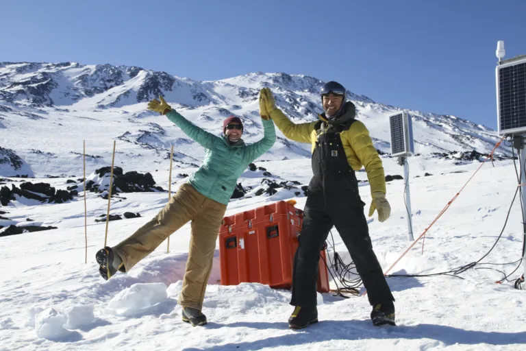 two people high-five in front of seismic instrument case and solar panels in the snow with mountain in background